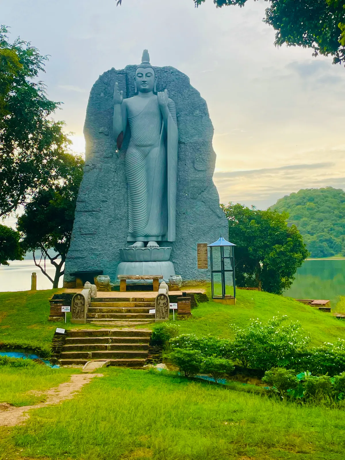 A tall standing Buddha statue on a green lawn by the lake at Giritale